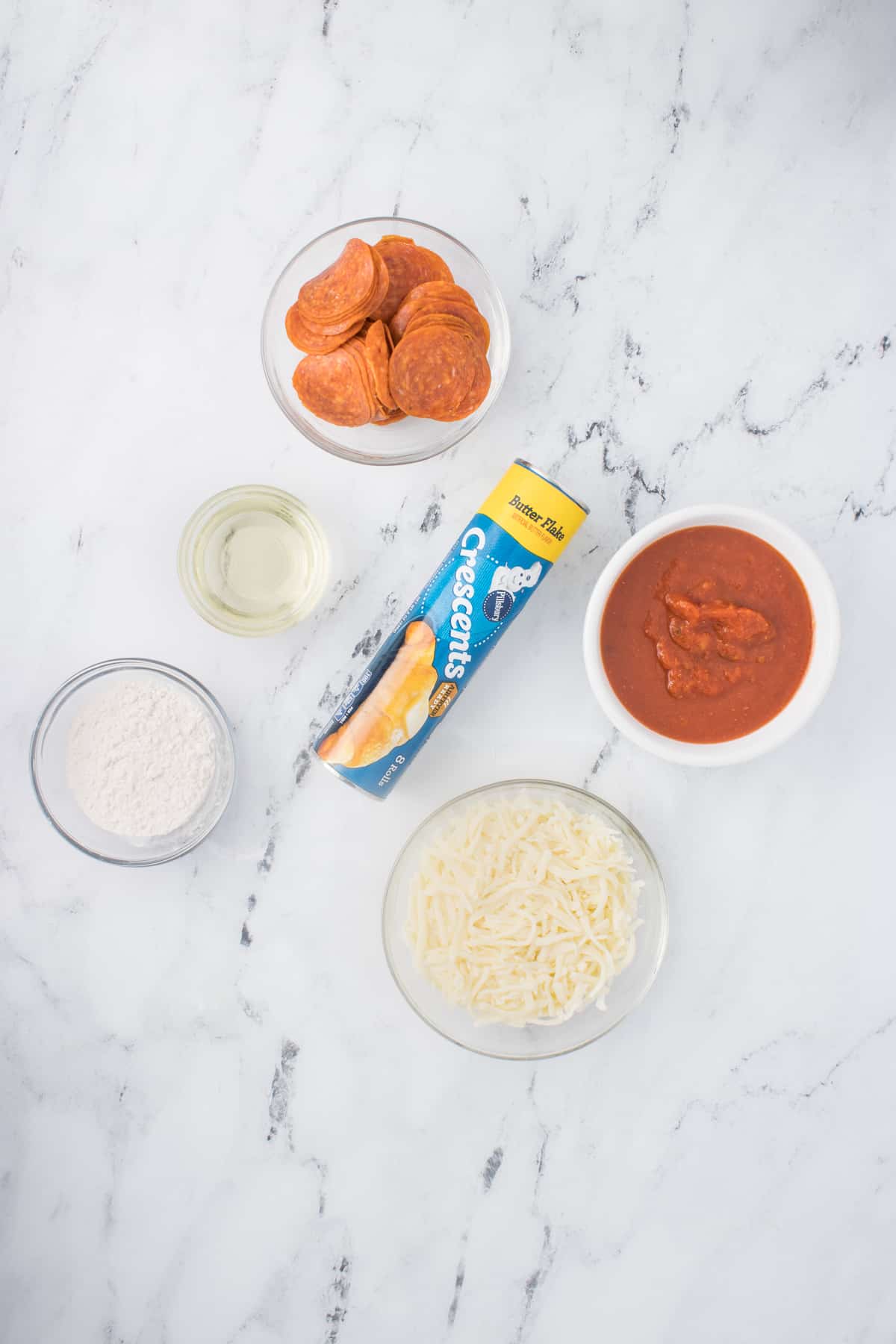 In this image, you can see a collection of ingredients ready to be used. The ingredients include crescent roll dough, shredded mozzarella cheese, pizza sauce, chopped pepperoni slices, all-purpose flour, and oil. 
