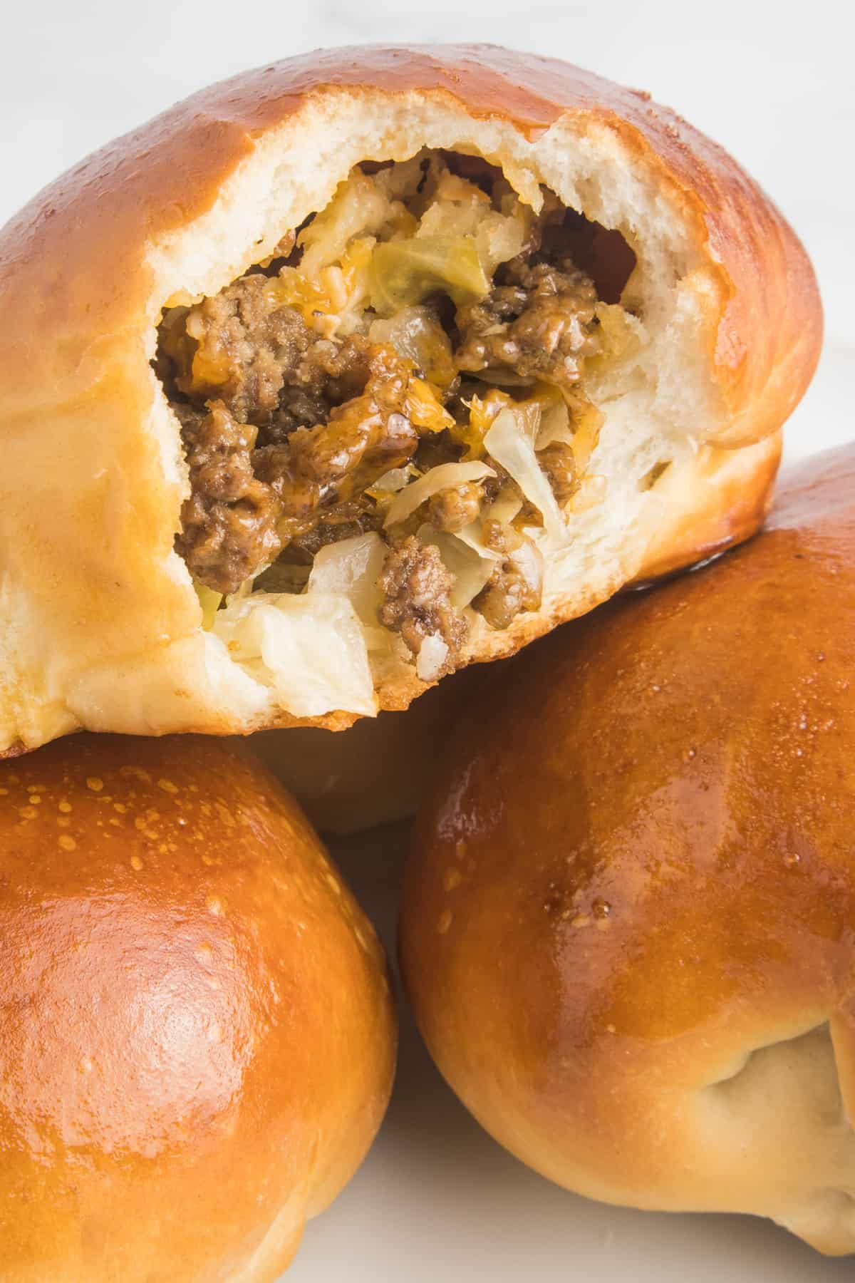 This image showcases a close-up shot of a runza, with a bite taken out of it, revealing the delicious contents inside. The runza is filled with meat, cheese, and shreaded cabbbage. It is placed on top of two unbitten runzas, forming a stack.