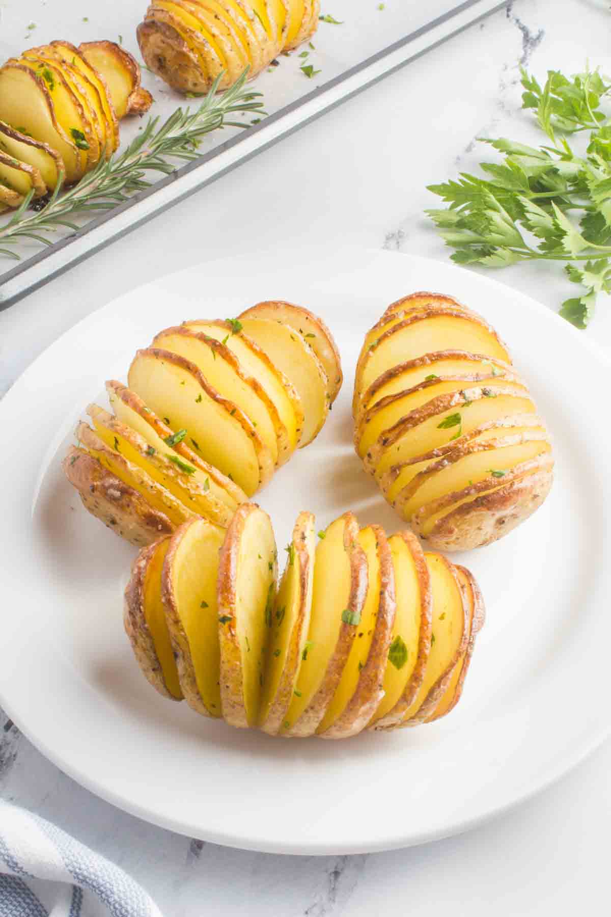 The image displays three rosemary hasselback potatoes on a plate. These potatoes have been sliced and seasoned, with rosemary adding a fragrant touch. Next to the plate, there is a baking pan containing the remaining cooked potatoes, ready to be enjoyed.