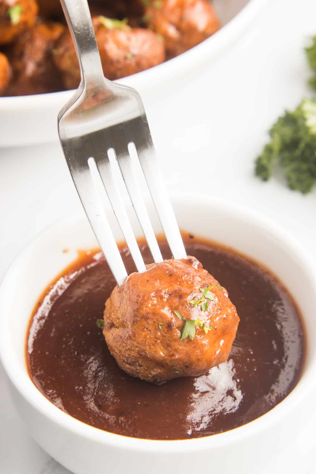 The image captures a single meatball delicately perched on a fork, glistening with sauce as it is being dipped, with the sauce coating the meatball.