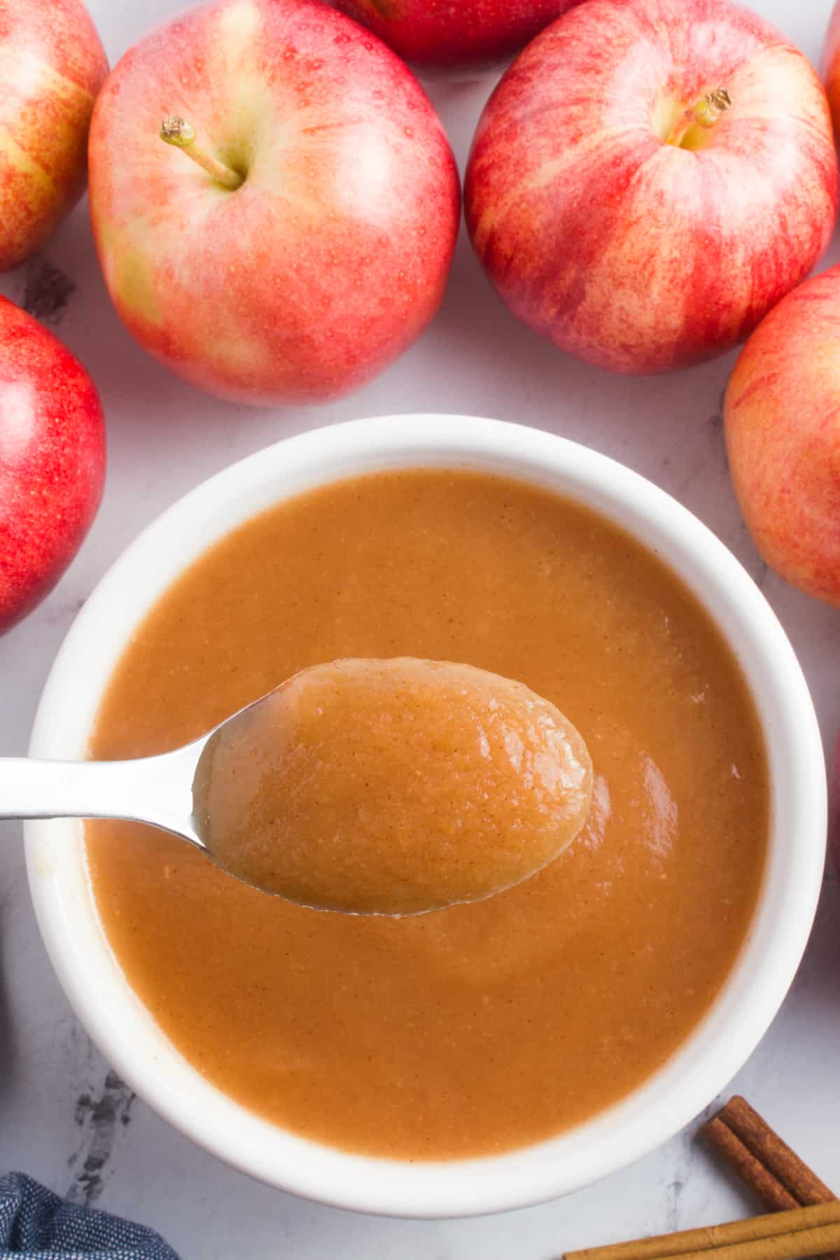 A spoon of applesauce is shown in the foreground, its smooth and velvety texture on full display. Behind the spoon, a bowl of applesauce is visible, suggesting the abundance of this delicious treat. Red apples are positioned around the bowl, adding a touch of color and visual appeal. Two cinnamon sticks are placed nearby, suggesting the aromatic flavor that complements the applesauce.