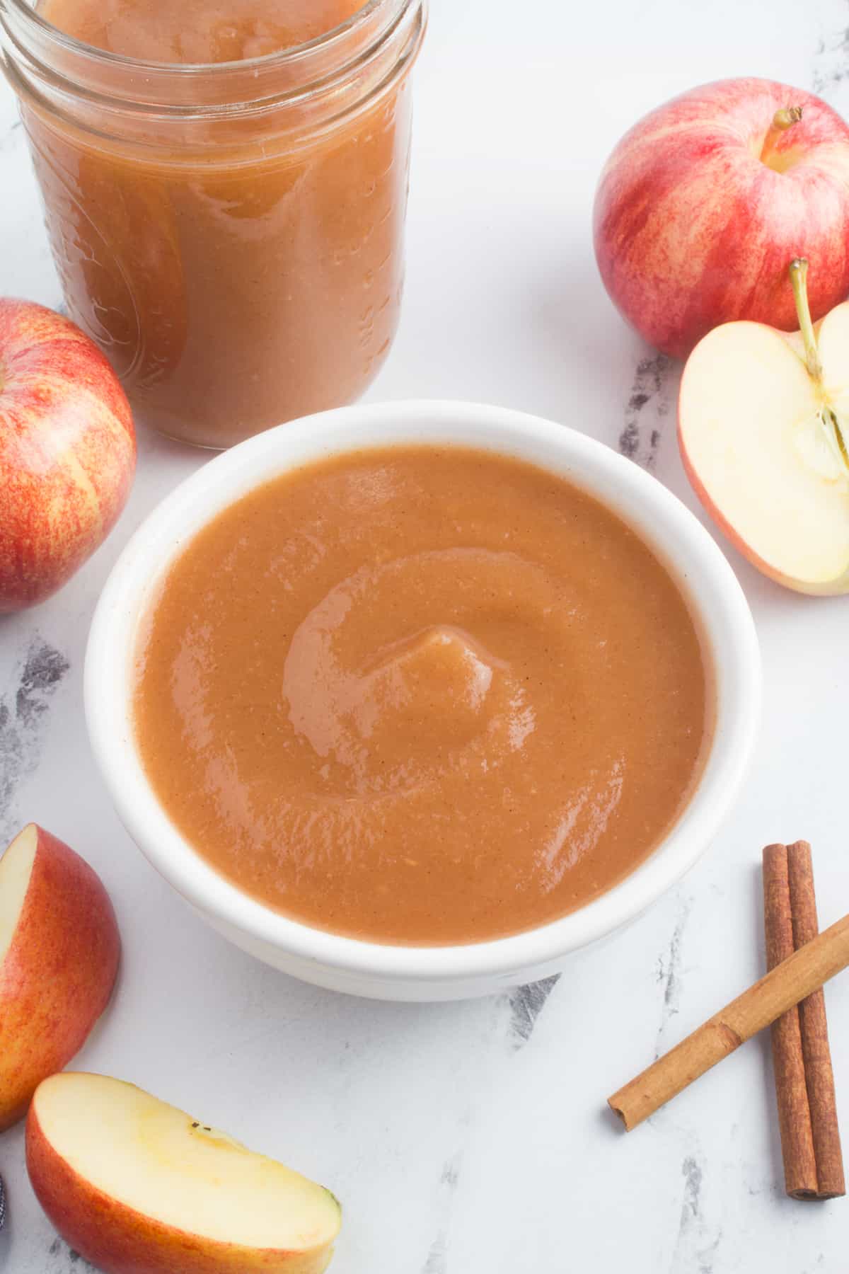 A bowl of freshly made applesauce is the centerpiece of the image. Red apples surround the bowl, and two cinnamon sticks rest nearby. A mason jar filled with more applesauce suggests that there is plenty of this delicious homemade treat to go around.
