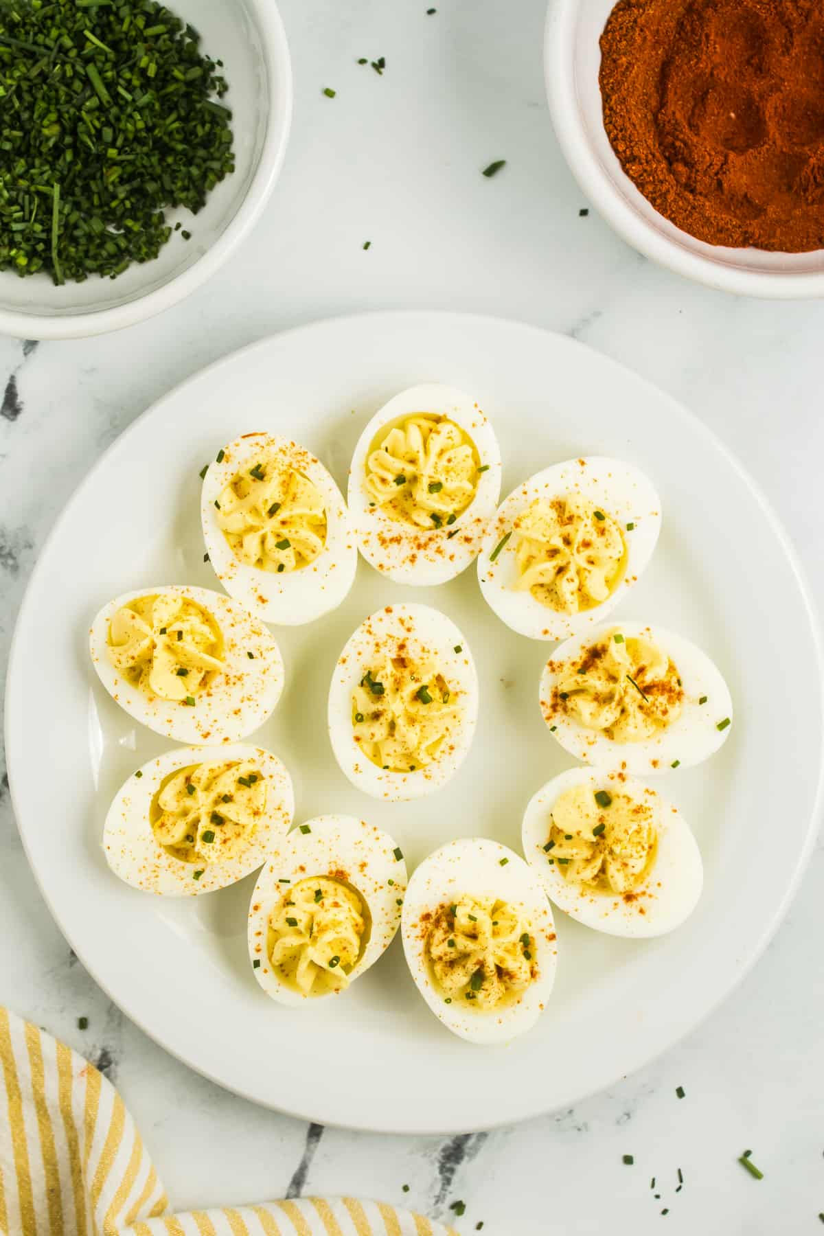 The image features a plate of Deviled Eggs with Apple Cider Vinegar. The deviled eggs are arranged neatly, showcasing their appetizing appearance. On the side, there is a small bowl of paprika, adding a touch of color and flavor to the dish. Additionally, chopped chives are placed nearby, providing a fresh and aromatic garnish option.