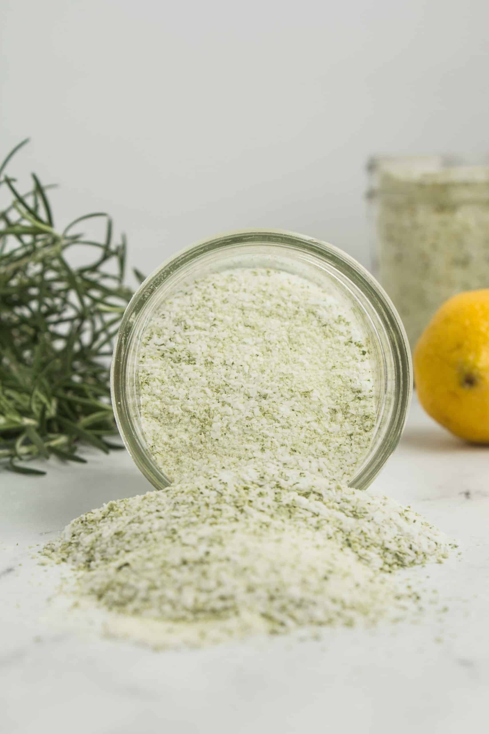 The image portrays a tipped-over mason jar with a heap of freshly made rosemary salt spilled in front of it. The rosemary salt is a flavorful blend, combining the aromatic essence of rosemary with salt.