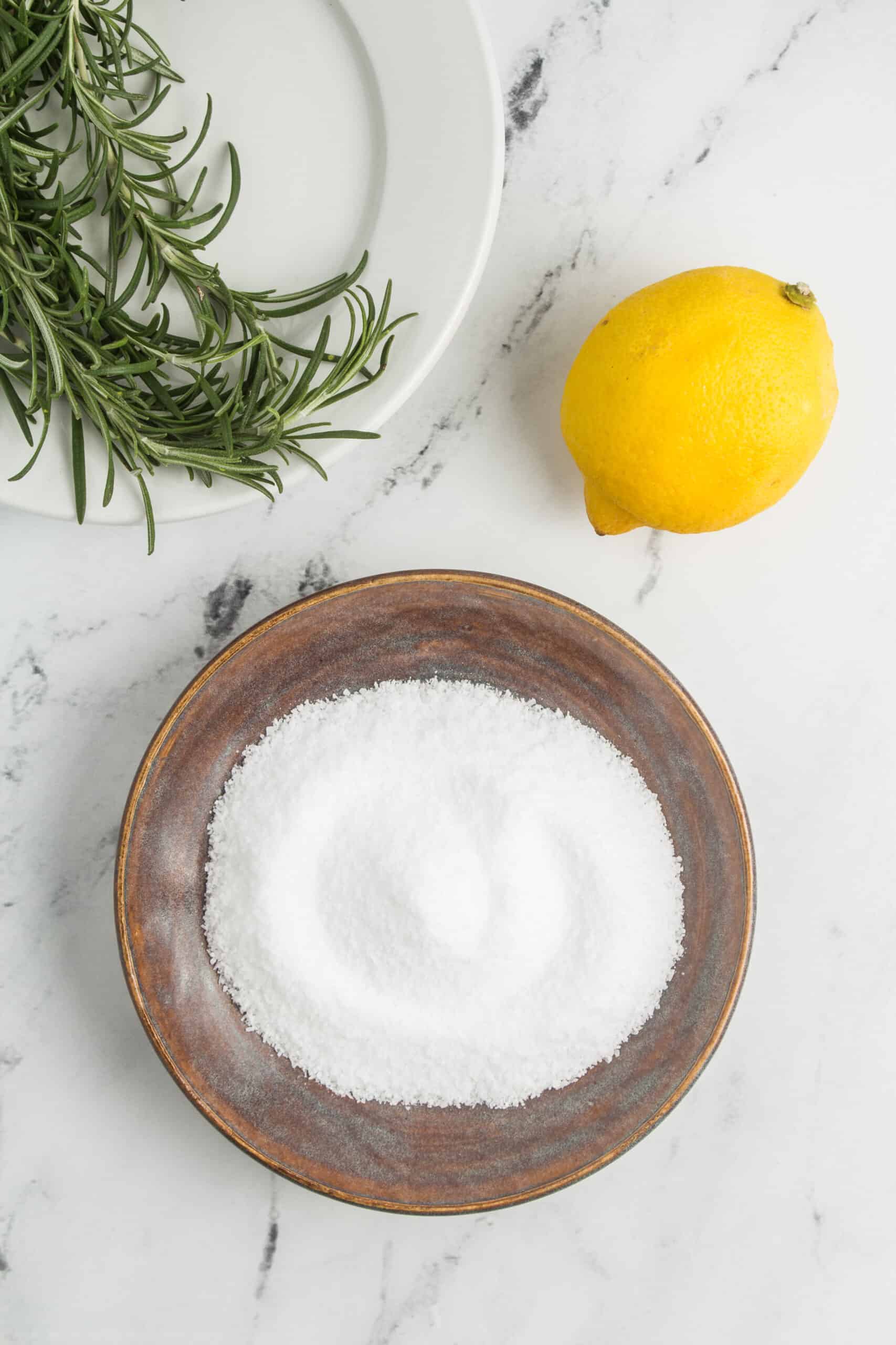 This ingredient shot features three key elements: salt, fresh rosemary, and a lemon. The salt is seen in a container, providing the base for the recipe. Next to it, a bunch of fresh rosemary sprigs is displayed, exuding a fragrant aroma. Lastly, a lemon is included, showcasing its bright and citrusy presence in the recipe.