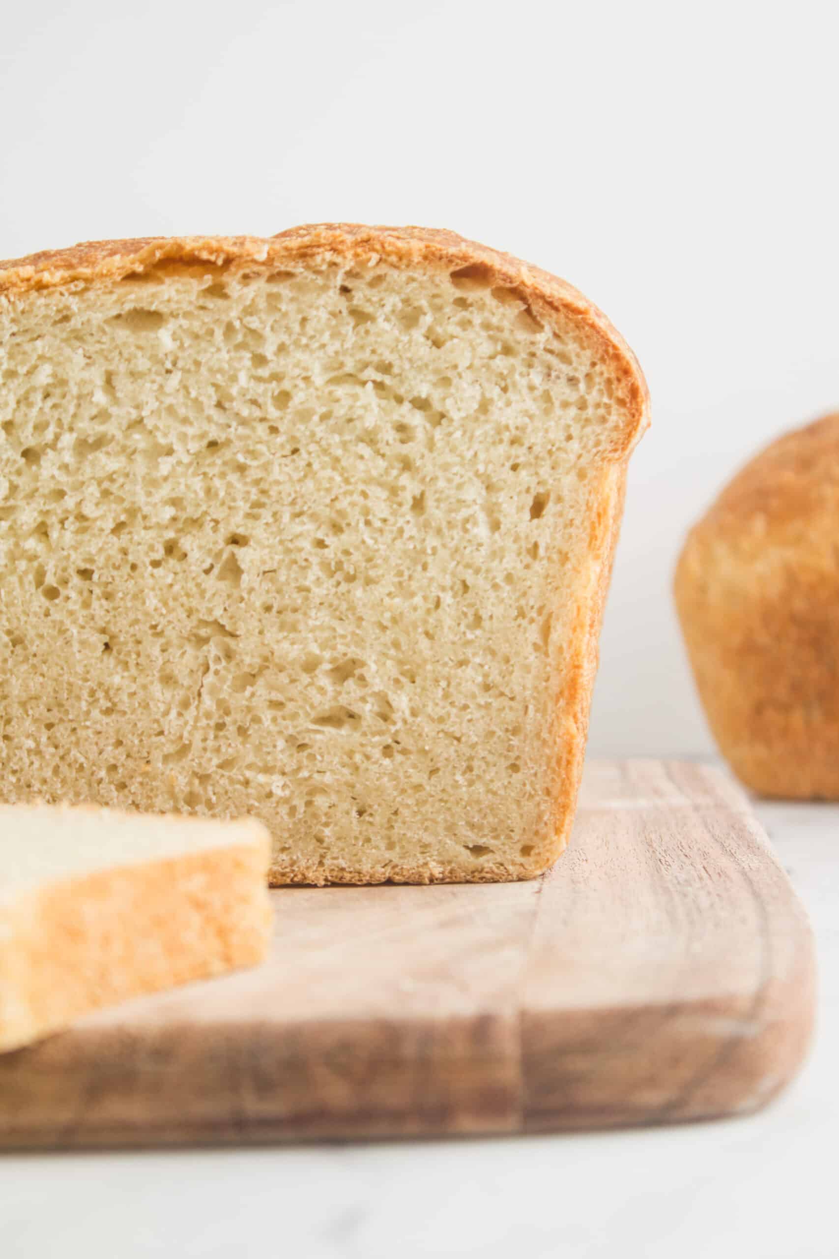 This image showcases a close-up "crumb shot" of sandwich bread, freshly cut and placed on a cutting board. Beside the loaf, there is a single slice of bread lying on its side. In the background, another loaf of bread can be seen, adding depth to the composition.