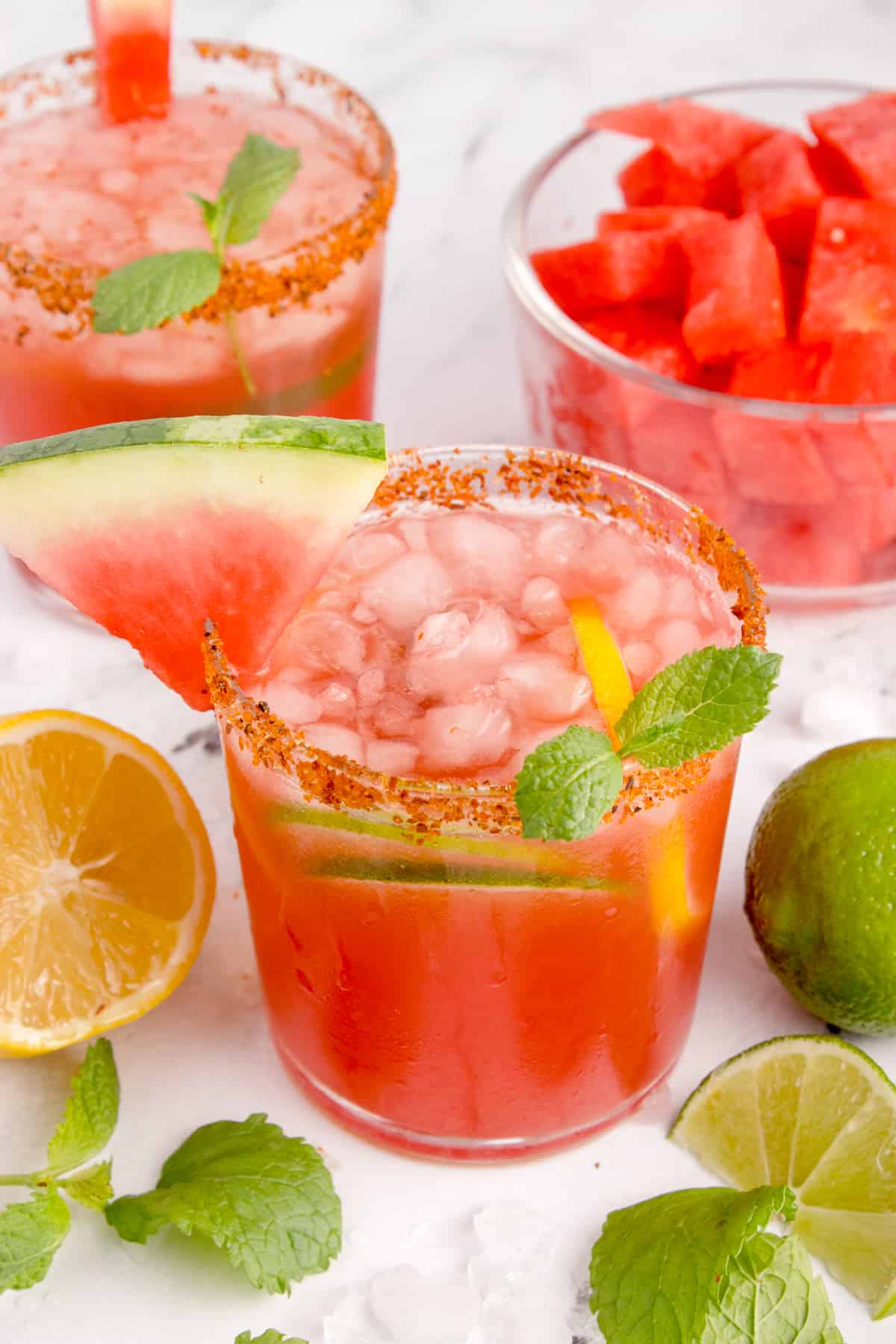 The image displays 2 glasses filled with refreshing watermelon kombucha. There is crushed ice in the glass, a lemon wedge, a watermelon wedge and mint leaves. The glass is rimmed with delicious Tajin. There is also a bowl with sliced watermelon in the background. The glass in the foreground is flanked by mint leaves and lemon and lime wedges. 