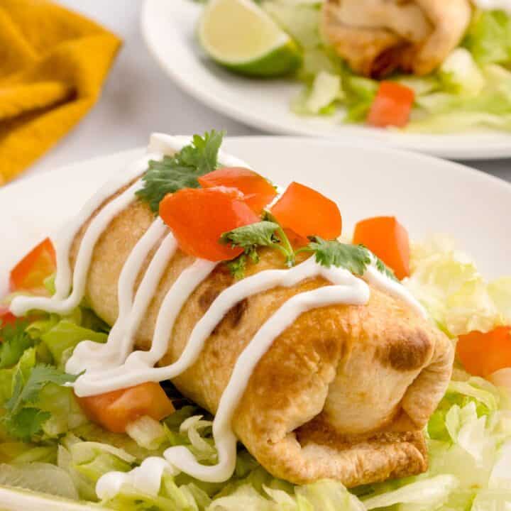 Image of mini chimichanga sitting on a bed of lettuce and garnished with sour cream, cilantro, and cubed tomatoes.
