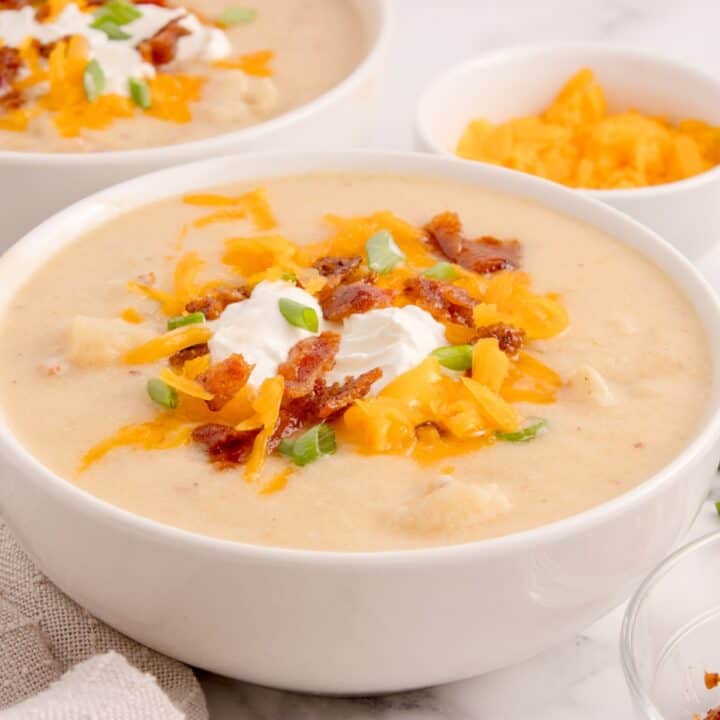 Cheesy potato soup in bowl, garnished with green onion, bacon, grated cheese and sour cream. Flanked by another bowl of creamy potato soup and a little bowls with grated cheese.